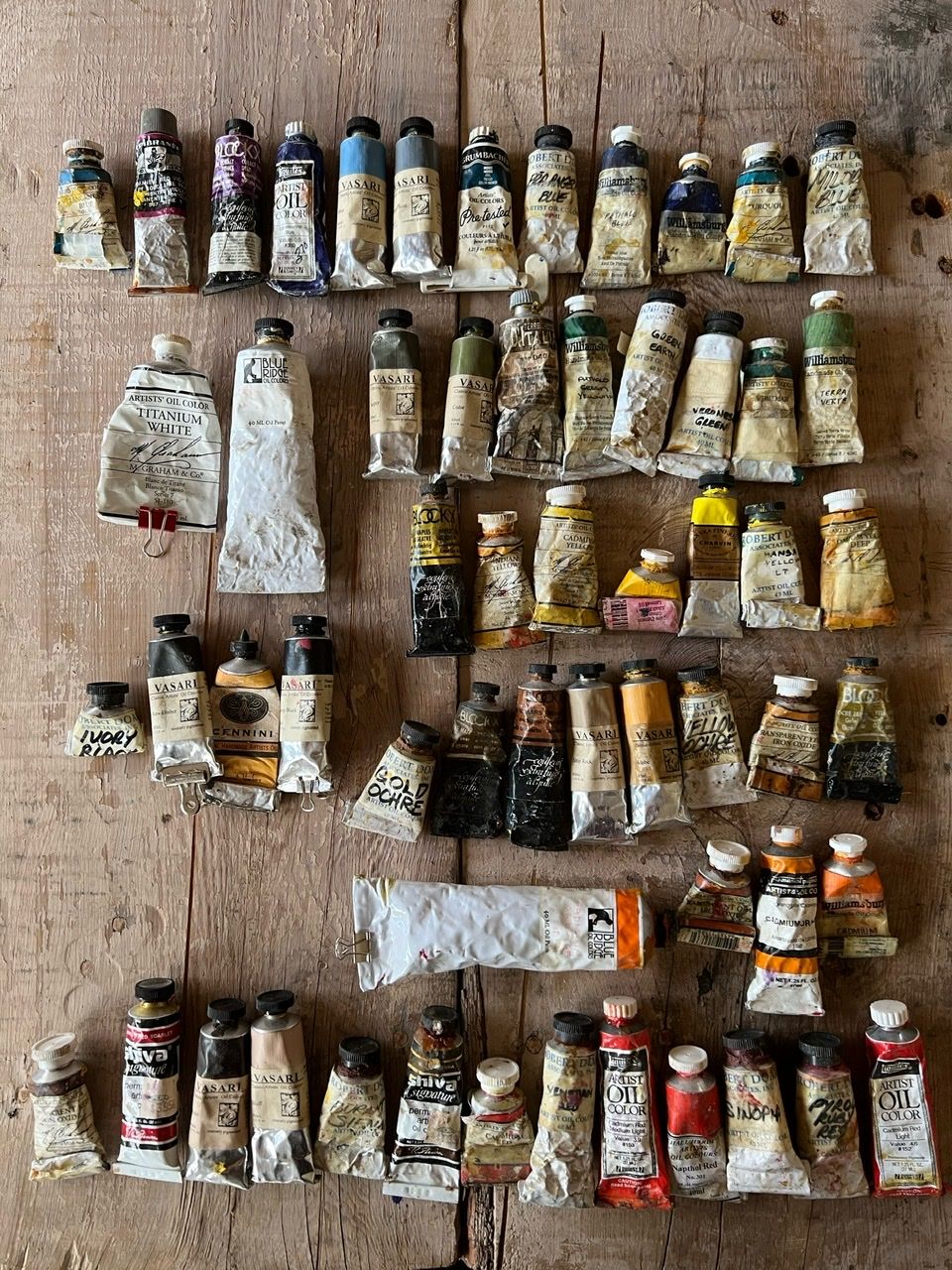 Overhead photograph of dozens of well-used oil paint tubes arranged in rows on a weathered wooden surface, featuring brands including Vasari, M. Graham, Blue Ridge, Robert Doak, and Williamsburg in a full spectrum of colors from titanium white to cadmium yellows, blues, greens, and deep reds.