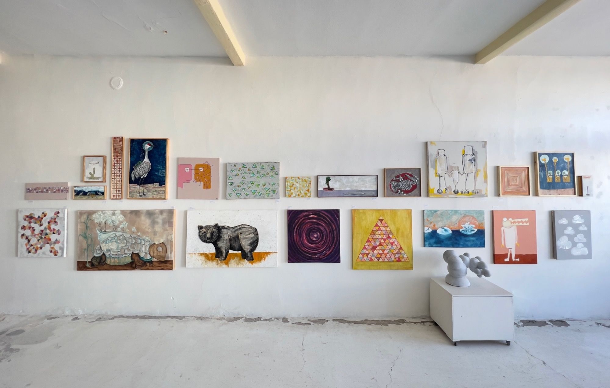 Gallery wall at Desert Archaic displaying two rows of paintings in varied sizes, including a large crane on deep blue, a bear, geometric triangles, abstract spirals, desert landscapes, robot-like figures, floral circles on pink, and a marble sculpture on a white pedestal at right