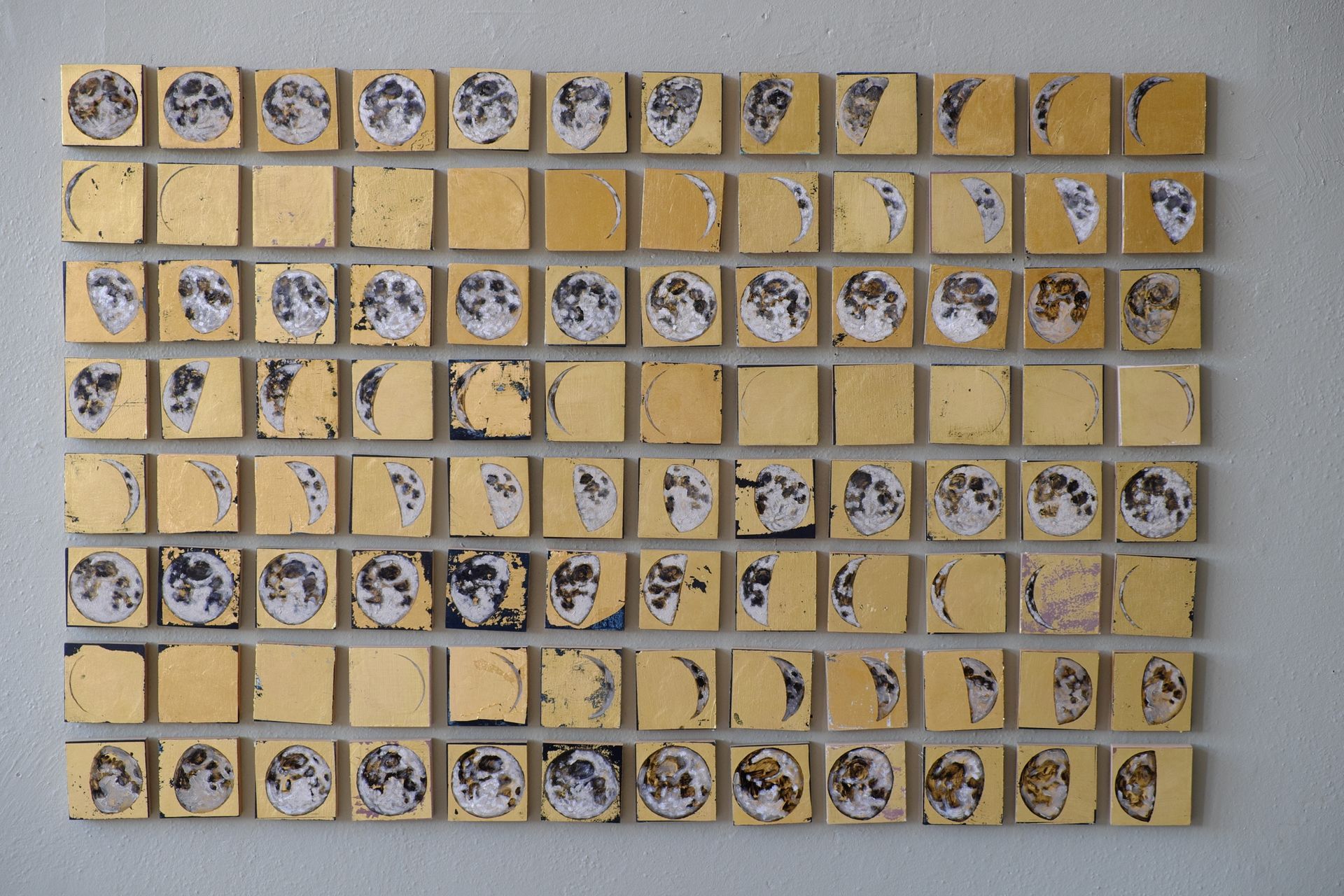 Frontal view of the complete Cyclum Lunarem grid, approximately ninety small linen squares gilded with gold leaf and painted with moon phases in white, gray, and black, arranged in even rows against a pale gray wall