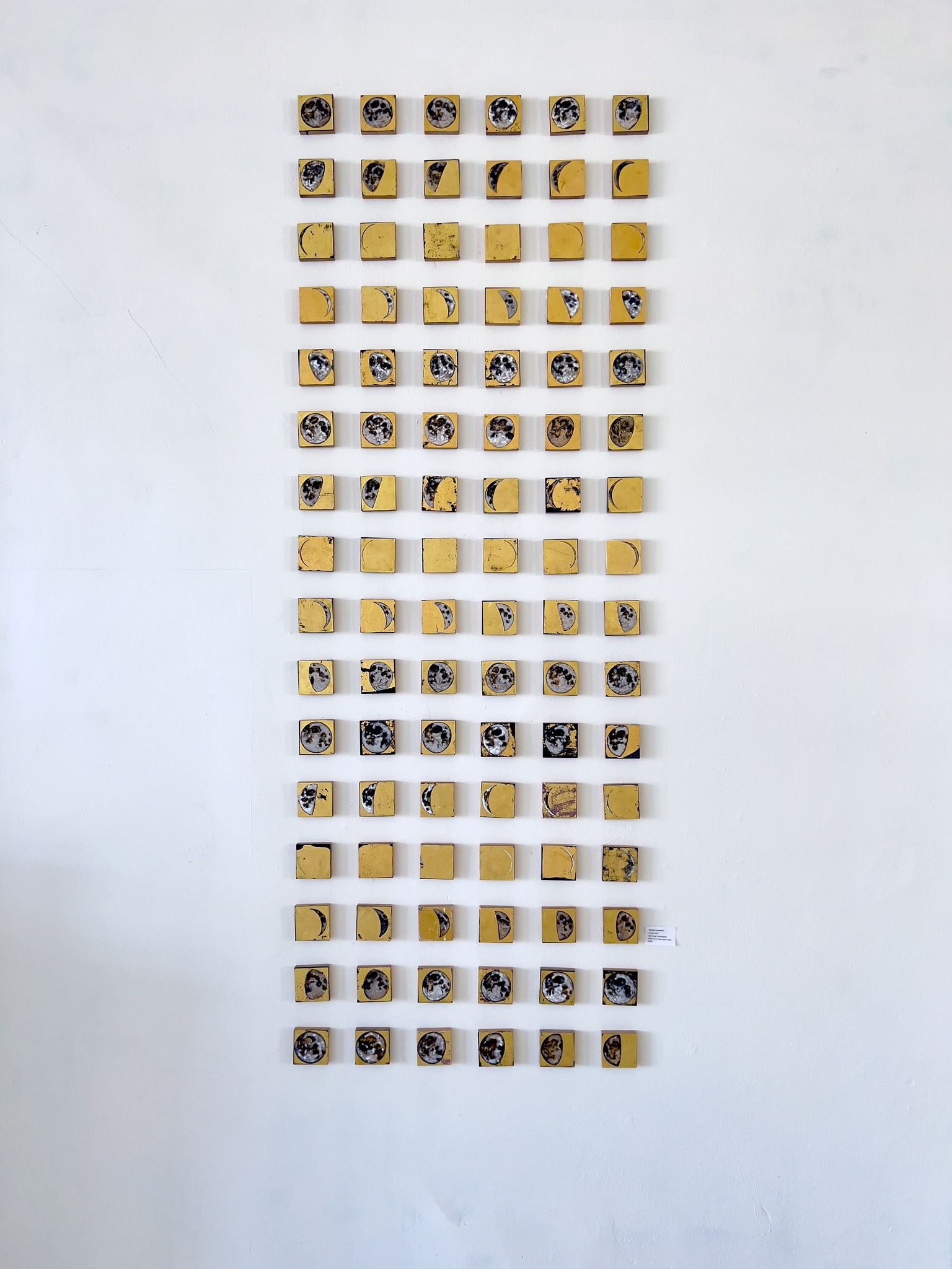 Cyclum Lunarem arranged in a tall vertical column on a white gallery wall, with rows of small gold-leafed squares depicting the full lunar cycle from new moon crescents to full moons in muted grays