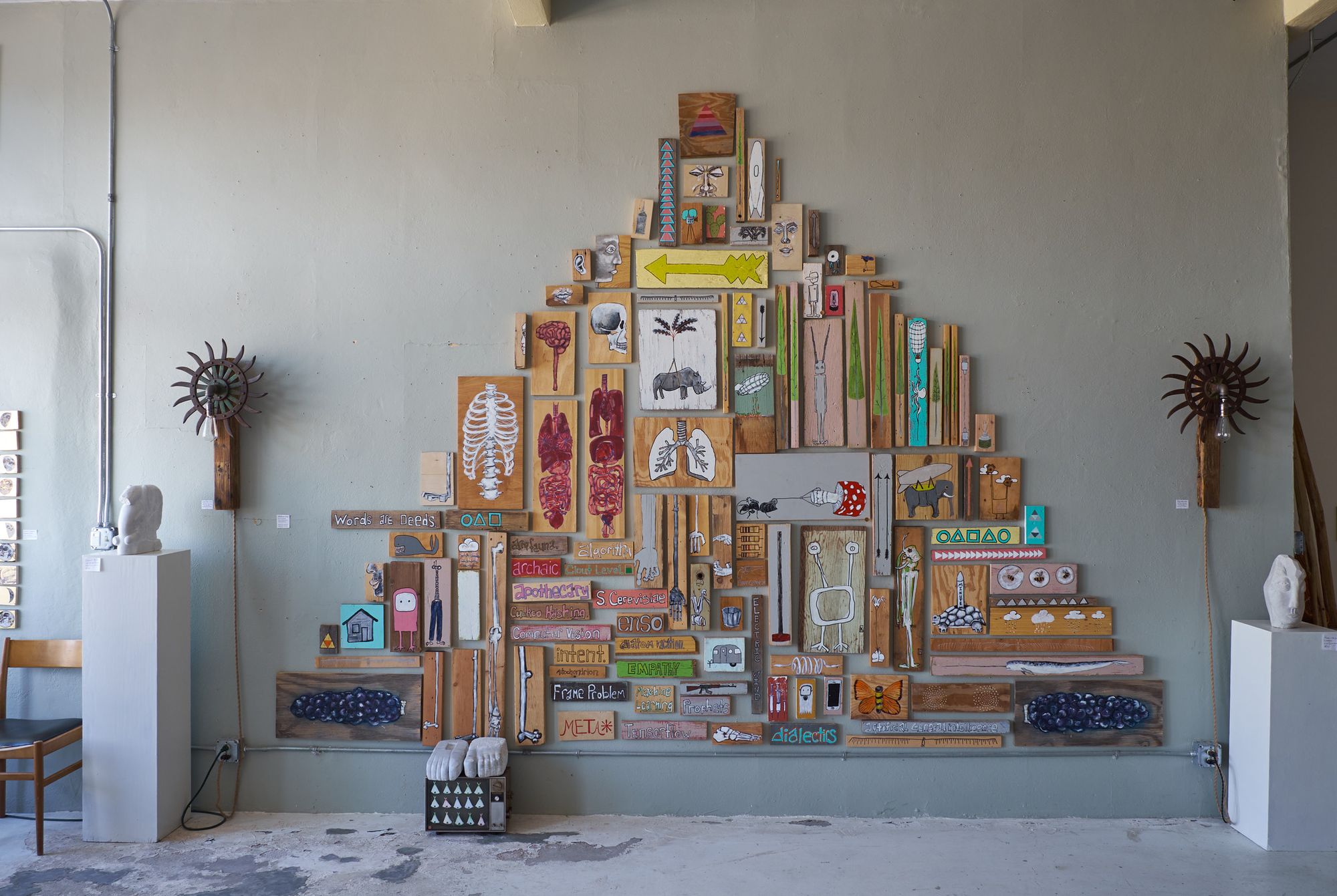 Pyramid-shaped wall installation at Desert Archaic gallery composed of dozens of painted found-wood panels and boards, featuring imagery of skulls, lungs, rib cages, animals, arrows, text, and geometric symbols in warm earth tones with colorful accents, flanked by two sun-shaped metal sculptures