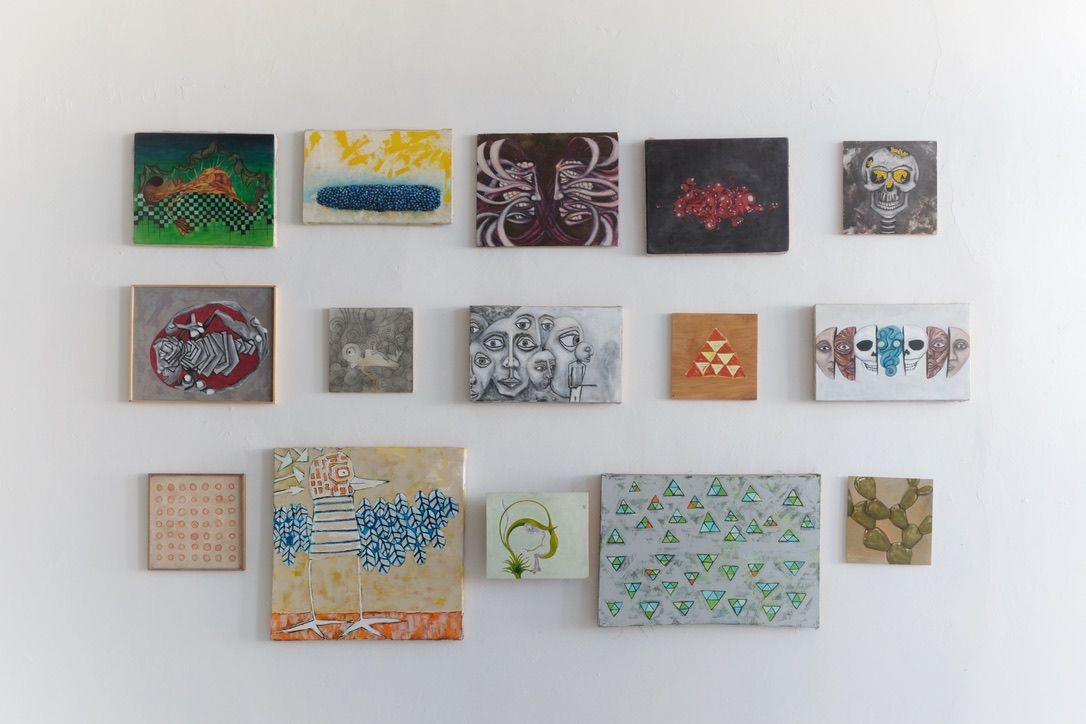 Fifteen small paintings arranged in a salon-style grid on a white wall, featuring diverse subjects including abstract faces, organic swirling forms, geometric triangles, skulls, figures, and botanical motifs in a range of greens, purples, oranges, grays, and earth tones