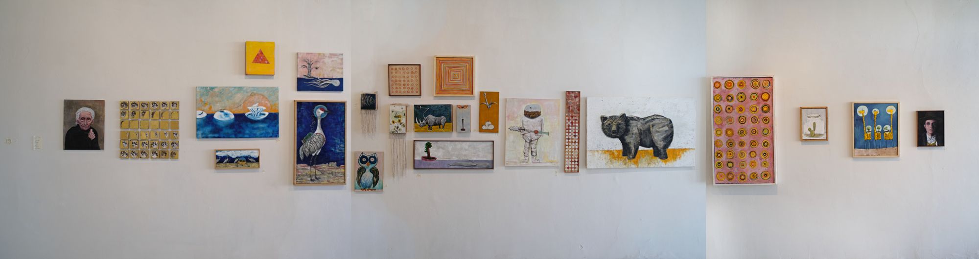 Wide gallery view of twenty-three paintings hung salon-style on a white wall, featuring a large crane, a bear, colorful geometric triangles, figurative portraits, landscapes, circular patterns on pink backgrounds, desert flora, and a grid of gold-leaf moon phases, with works ranging from small to large scale