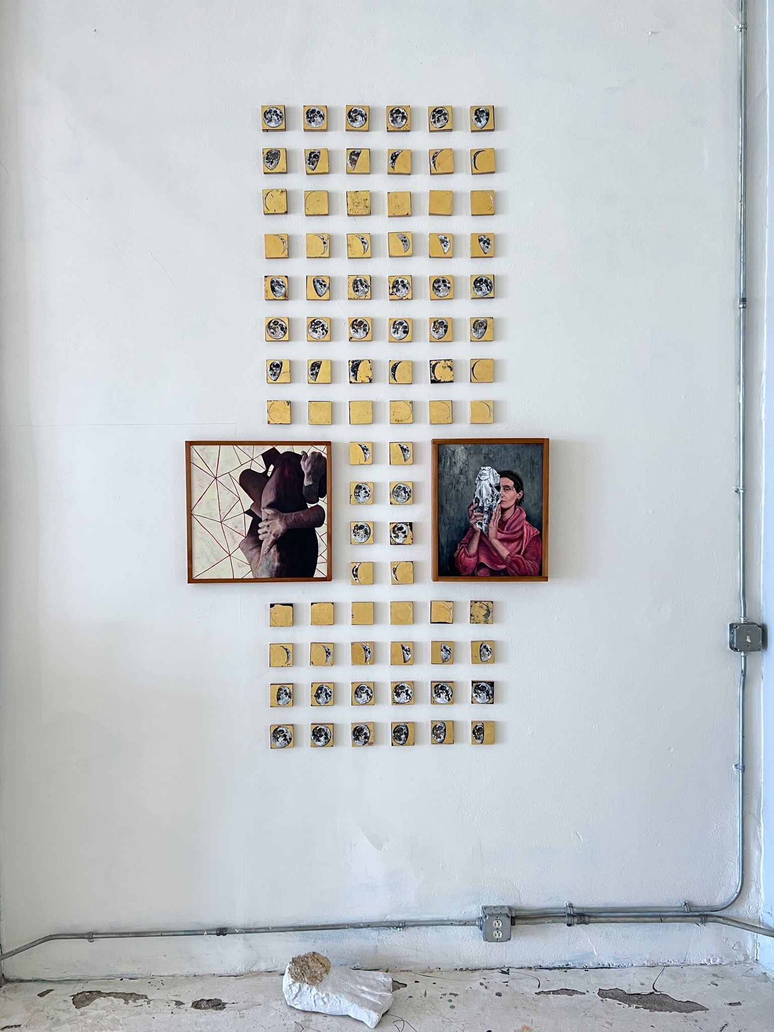 Installation combining Cyclum Lunarem gold-leaf moon phase grid with two framed paintings — one depicting a geometric figure study and the other a portrait of a woman holding a horse skull — all mounted on a white gallery wall with exposed conduit