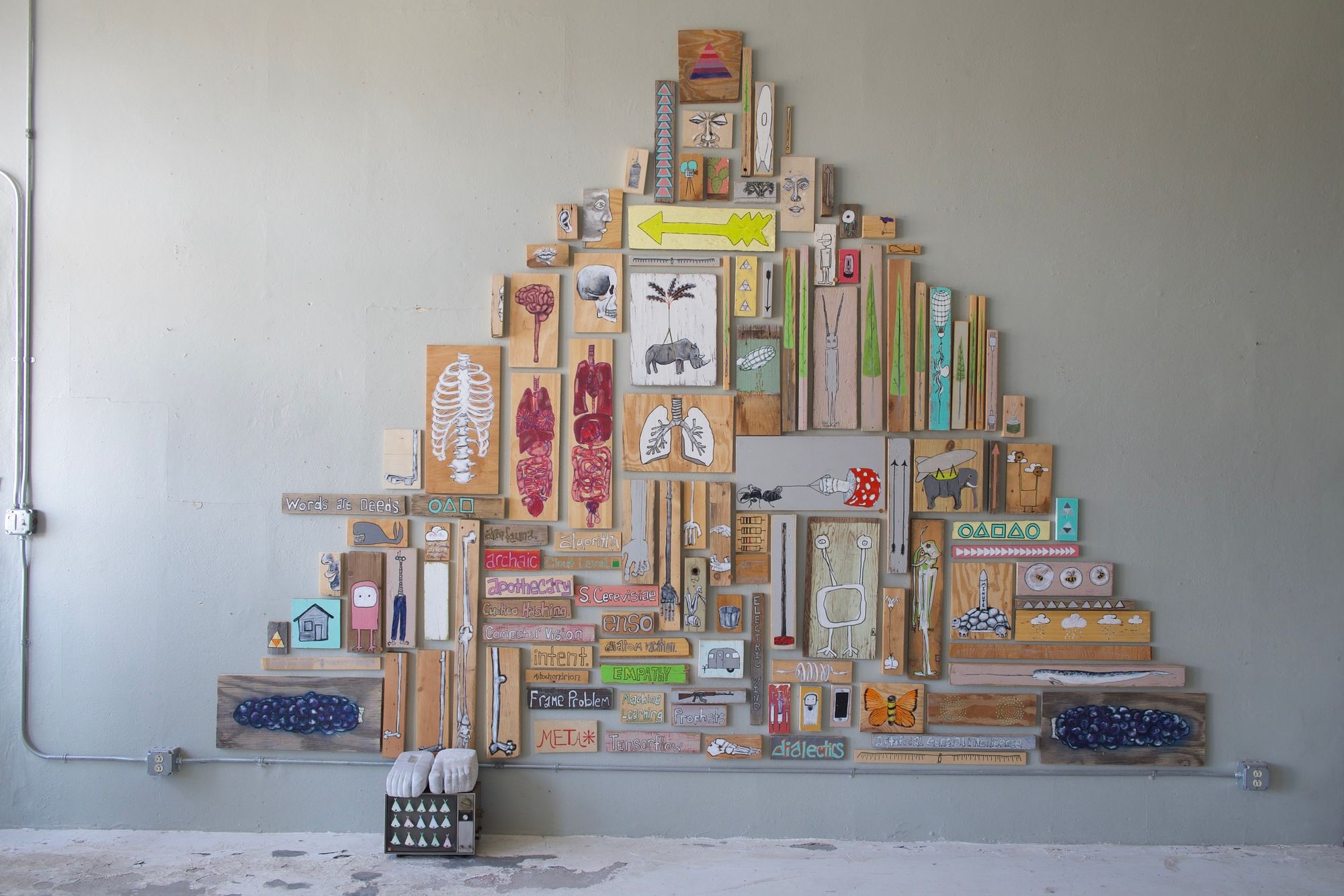 Wide view of a pyramid-shaped gallery installation composed of dozens of painted found wood panels featuring animals, anatomy, text, and whimsical imagery.