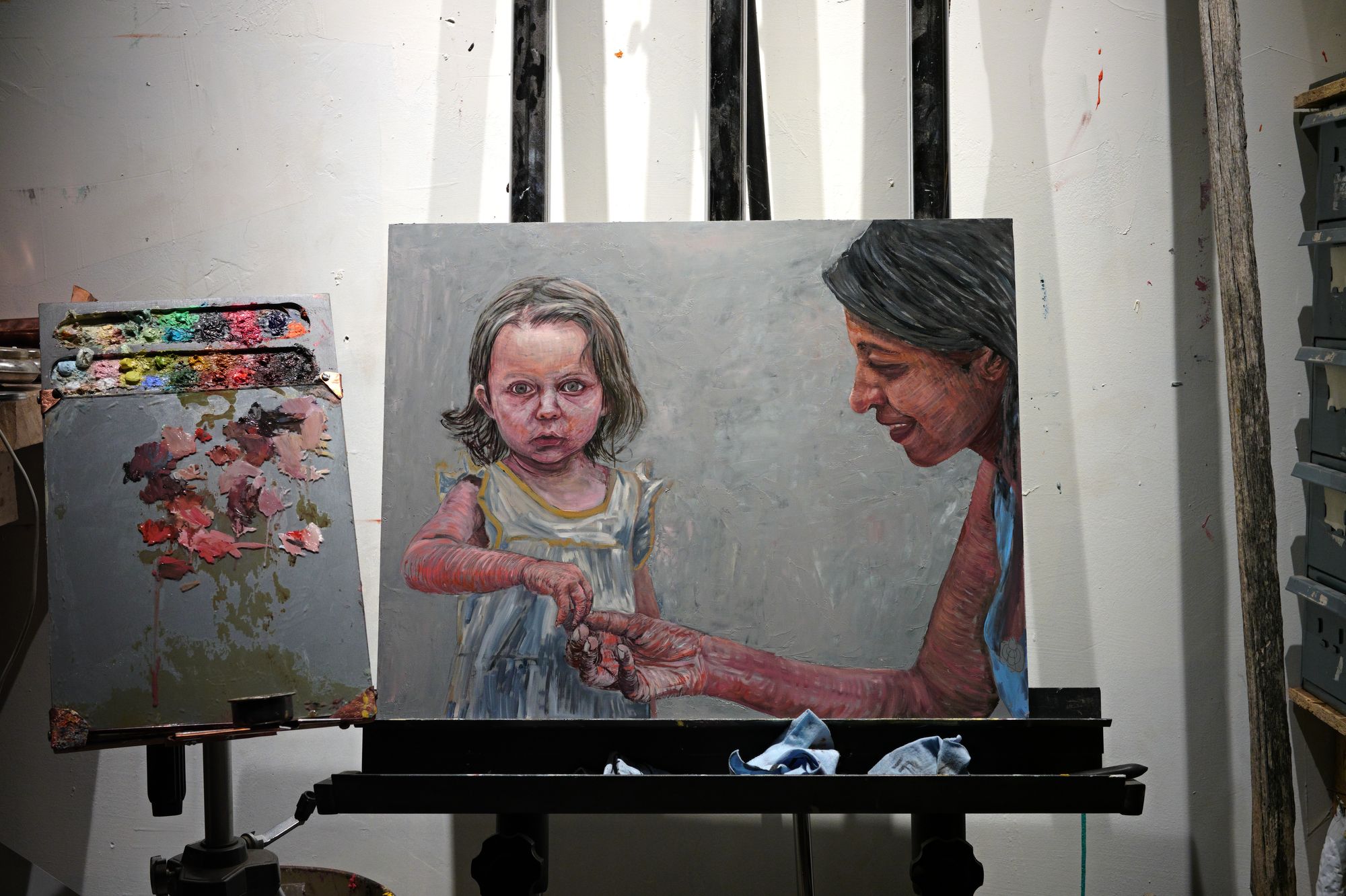 Oil painting in progress on an easel showing a young child in a white dress looking directly at the viewer while a woman beside her offers berries, rendered in expressive brushwork against a gray background.