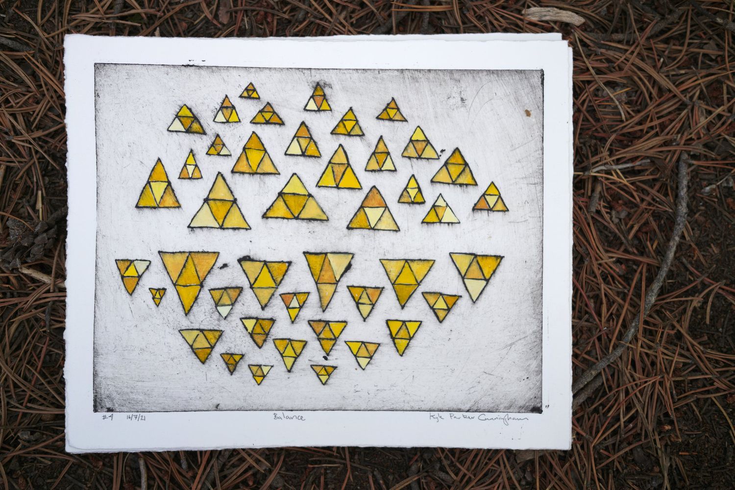 Drypoint intaglio print on Rives BFK paper featuring dozens of golden-yellow gouache-painted triangles in varying sizes scattered across the composition, each subdivided into smaller triangular shapes, photographed on a bed of brown pine needles.