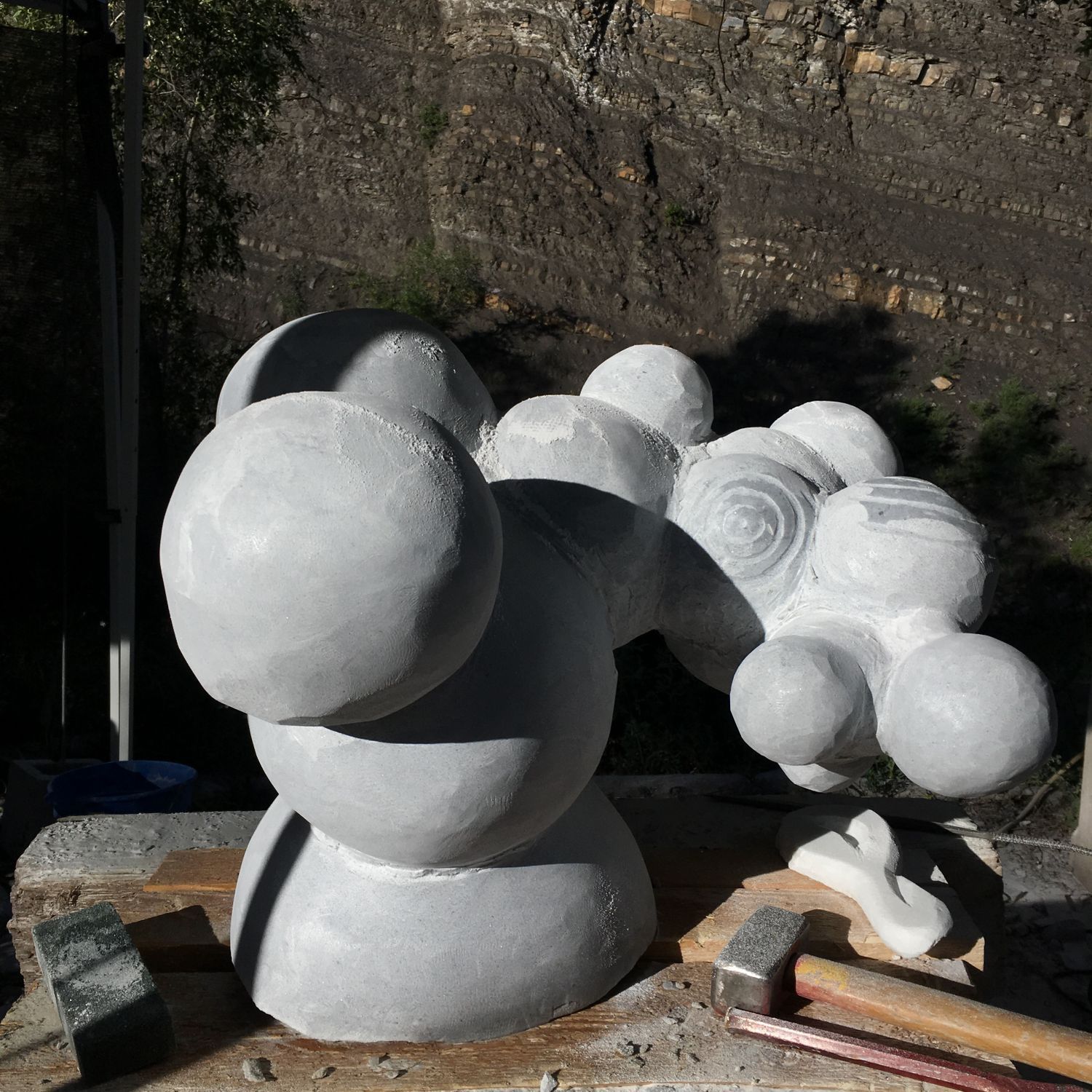 White marble sculpture in progress showing clustered spheres of varying sizes connected in a molecular form, sitting on a wooden workbench outdoors with carving tools and a stone wall in the background