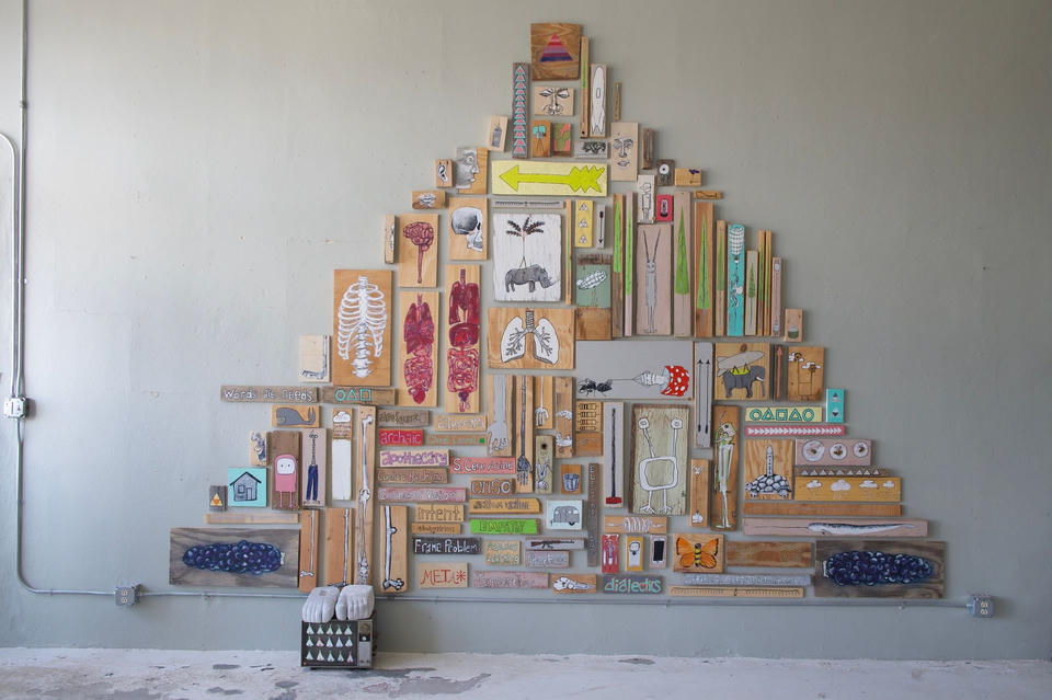 Wide view of a pyramid-shaped gallery installation composed of dozens of painted found wood panels featuring animals, anatomy, text, and whimsical imagery.
