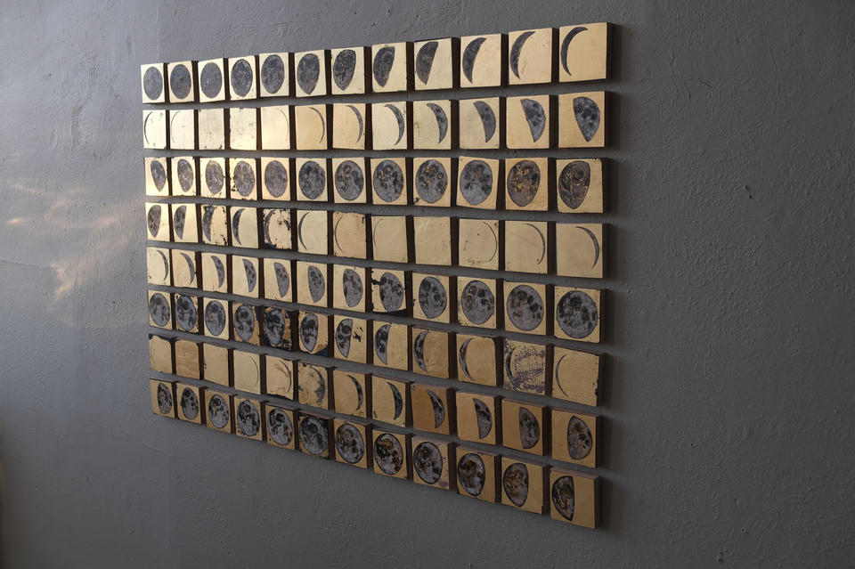 Angled view of Cyclum Lunarem showing rows of gold-leafed moon phase paintings catching atmospheric light, with silver and gray moons progressing from full to crescent across the grid