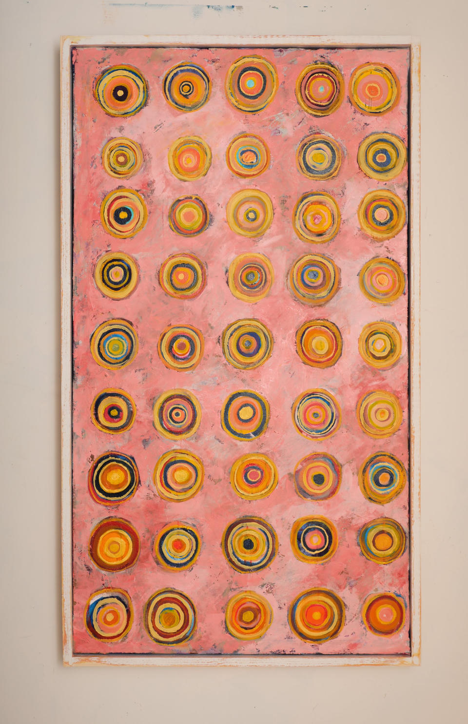 Oil painting featuring a grid of concentric circles in yellows, oranges, blues, and reds on a pink background, arranged in rows across a tall vertical composition.