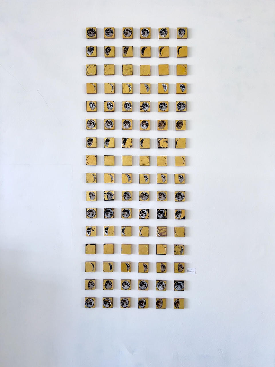 Full frontal view of Cyclum Lunarem installation arranged in a tall vertical column of gold-leafed square tiles, each painted with a lunar phase in muted grays and blacks on a white gallery wall