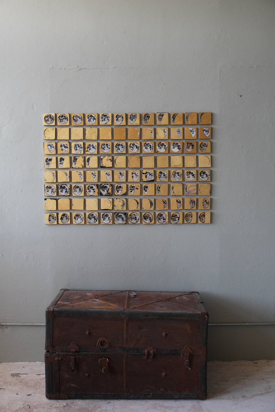 Grid of small square paintings on 24kt gold leaf depicting a summer's worth of moon phases, mounted on a gray wall above a vintage leather trunk