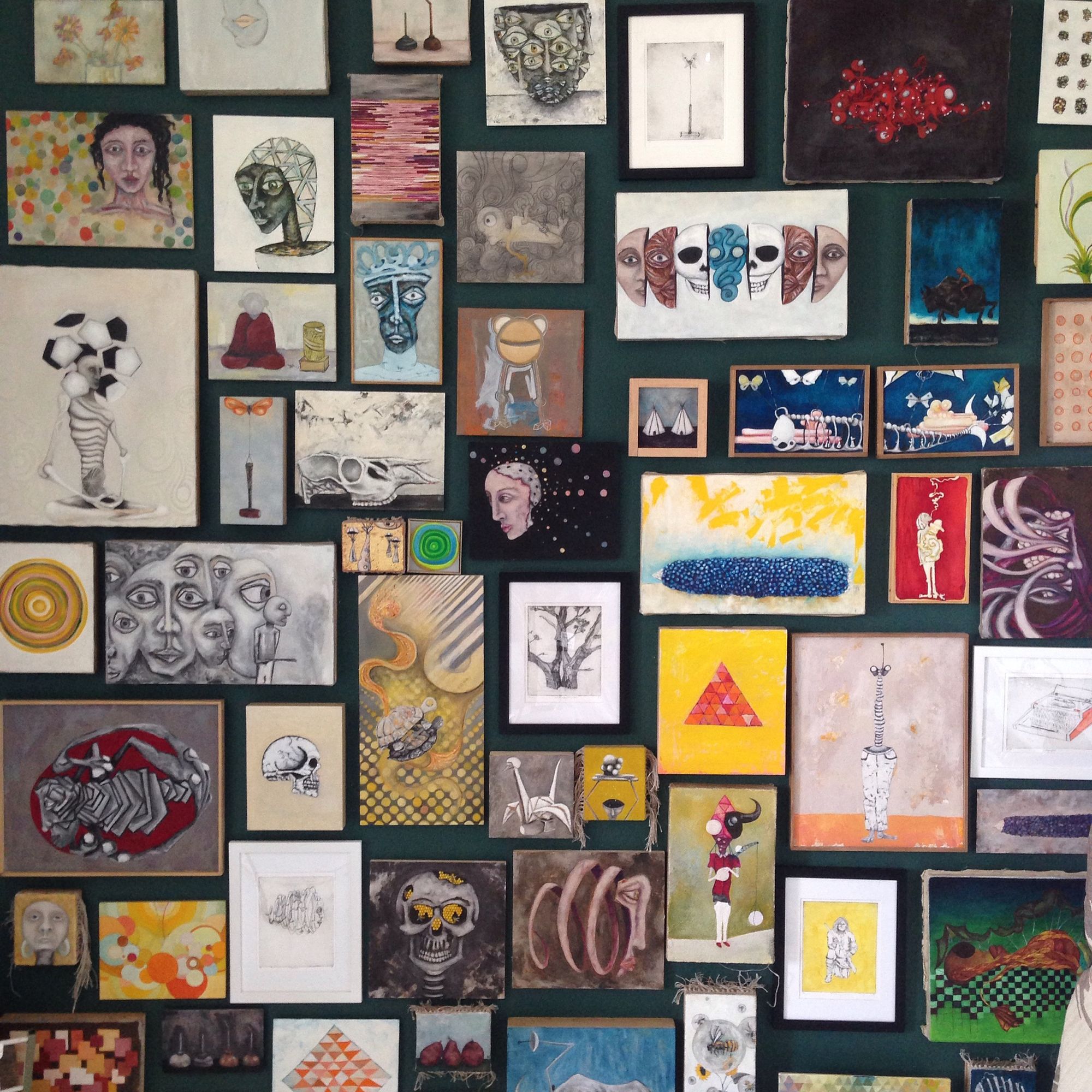 A dense salon-style wall display of dozens of small paintings and prints on a dark teal green wall, featuring portraits, still lifes, abstract forms, skulls, animals, and geometric shapes in a wide range of colors and styles.